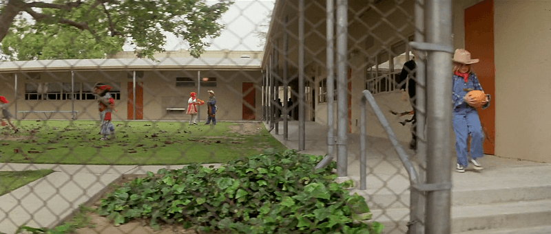 Uma cena de Halloween. A câmera está posicionada atrás de uma grade de ferro onde podemos ver uma escola com várias crianças usando fantasias de Halloween