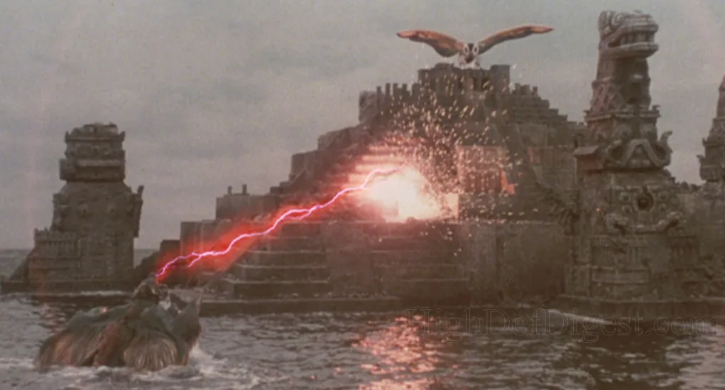 Cena do filme O Renascimento de Mothra 2. A titula Mothra está no topo de um templo antigo no meio do mar, enquanto um monstro marinho gigante a ataca com raios vermelhos