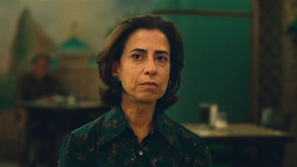 Na imagem tem Fernanda Torres interpretando Eunice Paiva no filme Ainda Estou Aqui. A câmera centra no rosto da personagem, que ela para longe, segurando uma lágrima no olho direito.
