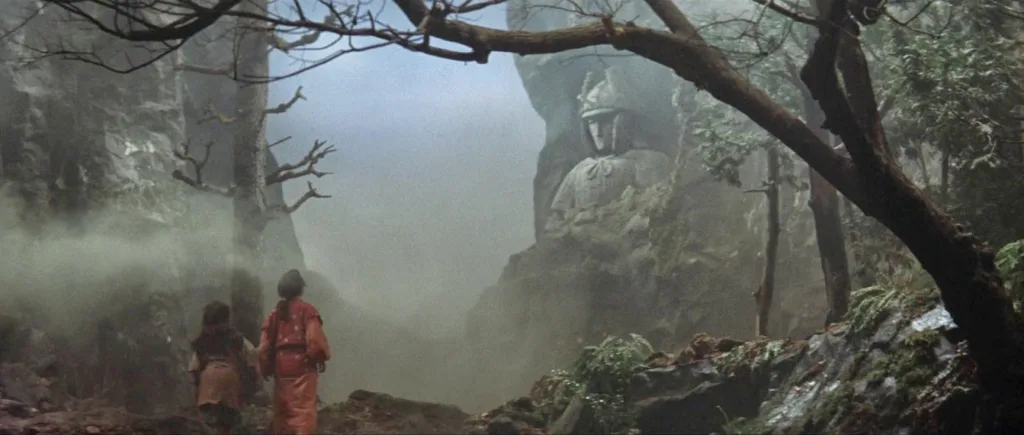 Cena do filme Daimaji. Duas pessoas, uma moça jovem vestindo um kimono vermelho, e uma criança com roupas desgastadas, estão numa montanha olhando para uma estátua gigante que retrata um guerreiro antigo, o Daimajin