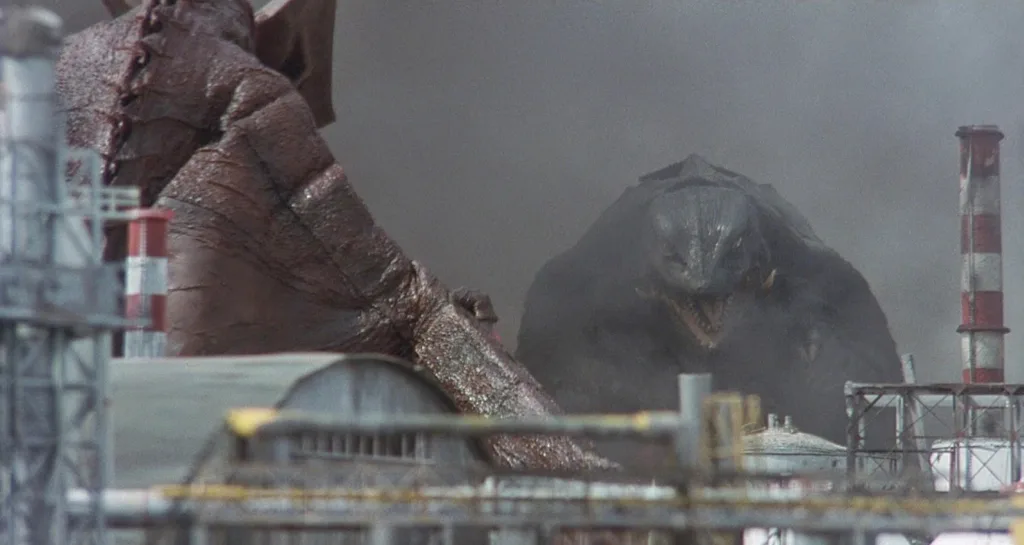 Cena do filme de Gamera: Guardião do Universo. A câmera filma o monstro Gyaos na sua forma final de costas, enquanto a frente dele Gamera aparece saindo de uma nuvem de fumaça. Os dois se se enfrentam num complexo industrial