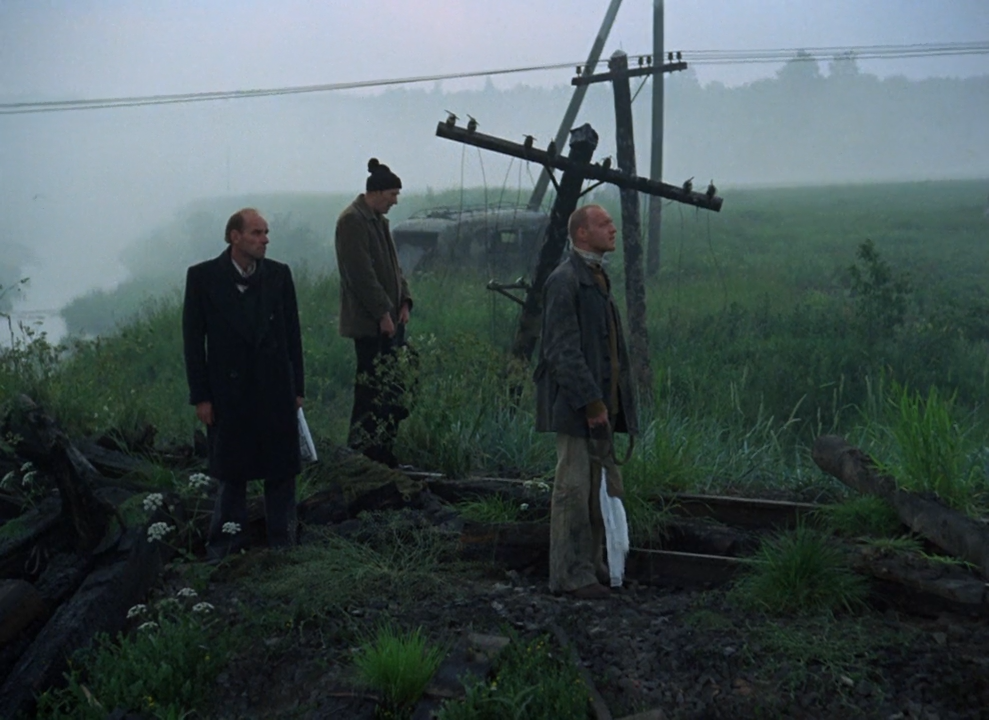 Cena do filme Stalker, mostrando os três personagens principais próximo a um trilho de trem numa paisagem desolada