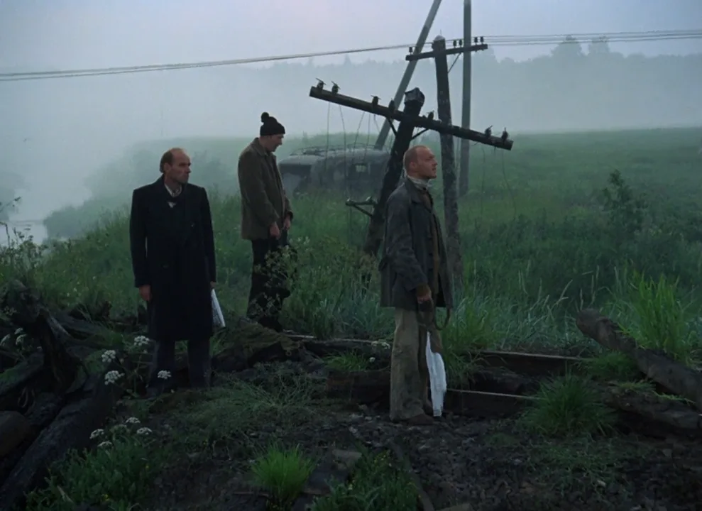 Cena do filme Stalker, mostrando os três personagens principais próximo a um trilho de trem numa paisagem desolada